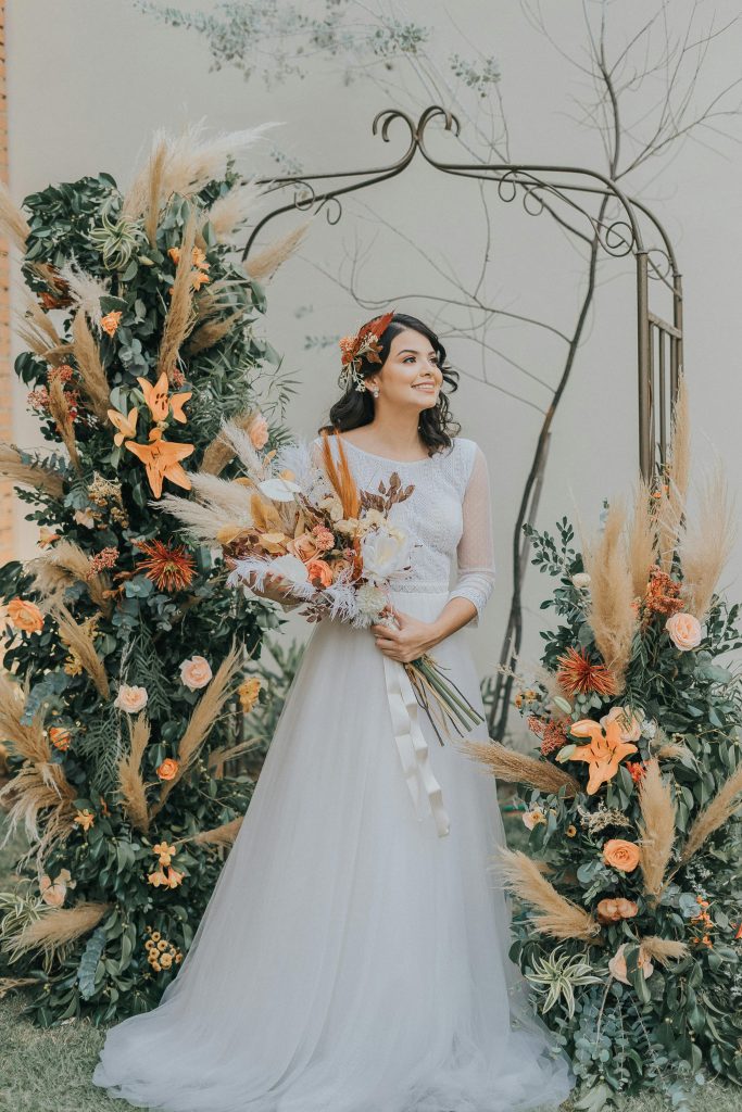 Bride in a graceful white dress surrounded by autumn-themed floral arrangements, smiling blissfully.
